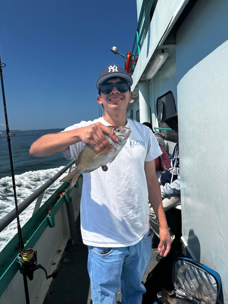 Person on a boat holding a fish, wearing sunglasses and a cap, with a fishing rod nearby.