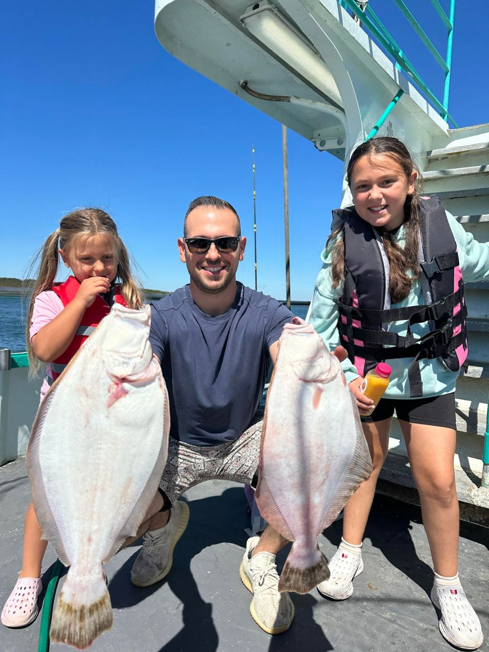 Man with two kids holding two large fish on a boat, all smiling under clear blue sky.
