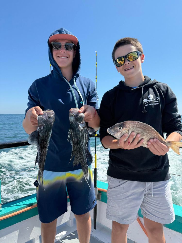 Two people on a boat holding fish, with ocean and clear sky in the background.