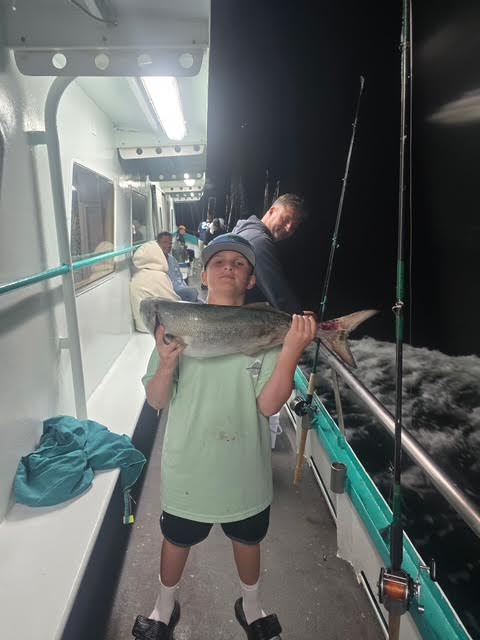 Child proudly holding a large fish on a boat at night, with others in the background.