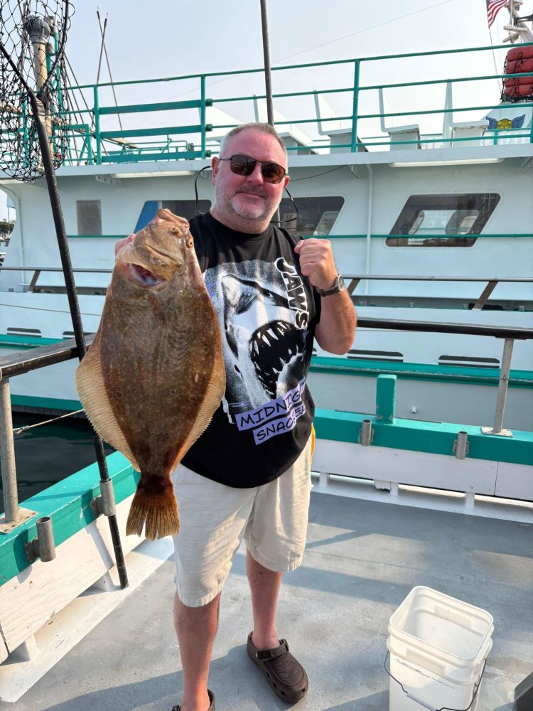 Man on boat holding a large flatfish, wearing a shark-themed t-shirt and sunglasses.