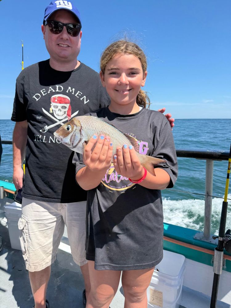 A man and girl on a boat. The girl is holding a fish, smiling. The ocean is visible in the background.