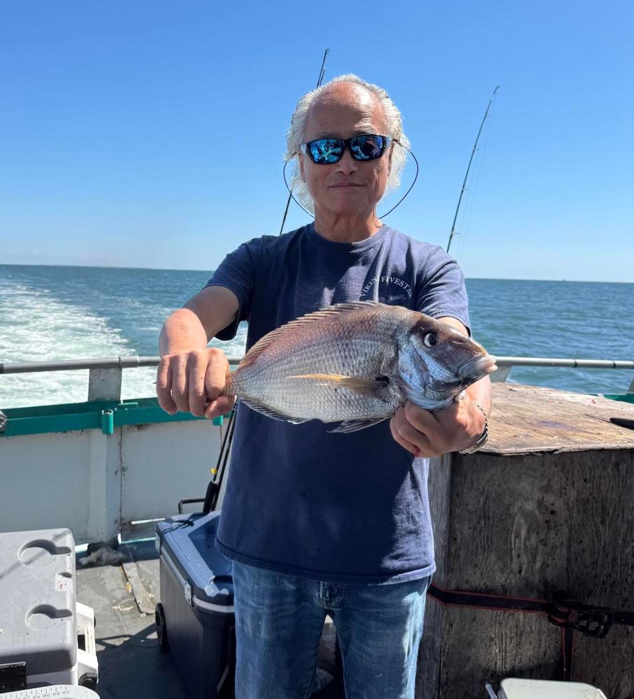 Person with sunglasses holding a large fish on a boat with ocean in the background.