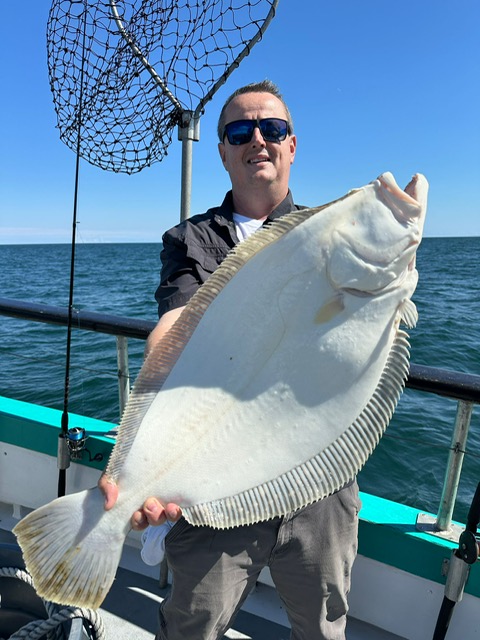 Person holding a large flatfish on a boat with the ocean in the background.