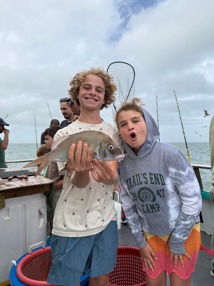 Two children on a boat holding a fish, with fishing gear and people in the background.