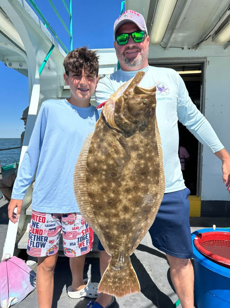 Two people on a boat holding a large flat fish, with a clear blue sky in the background.