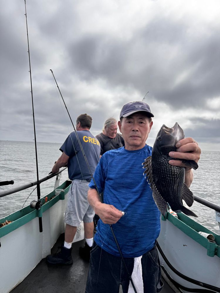 Man on boat holding fish, with two others fishing, under cloudy sky.
