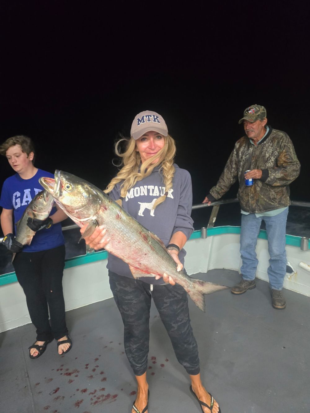 Person holding a large fish on a boat at night, with two others in the background.
