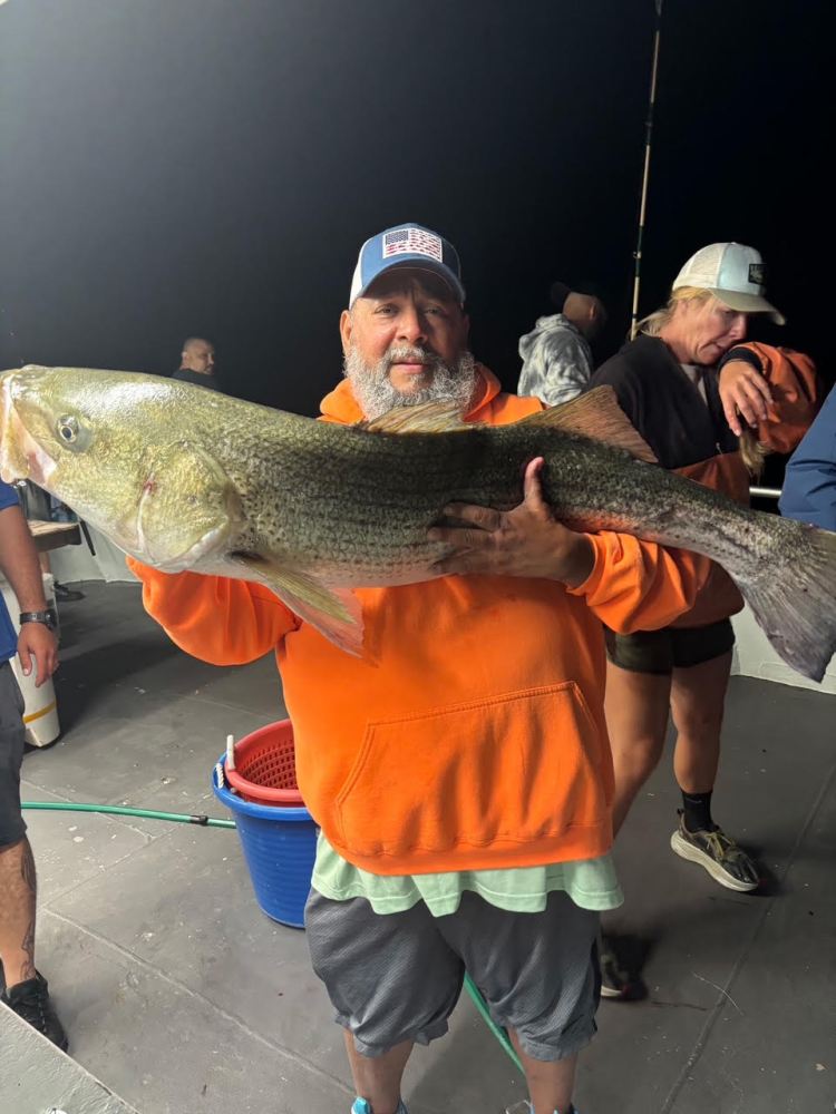 Person in orange hoodie proudly holding a large fish on a boat deck at night.