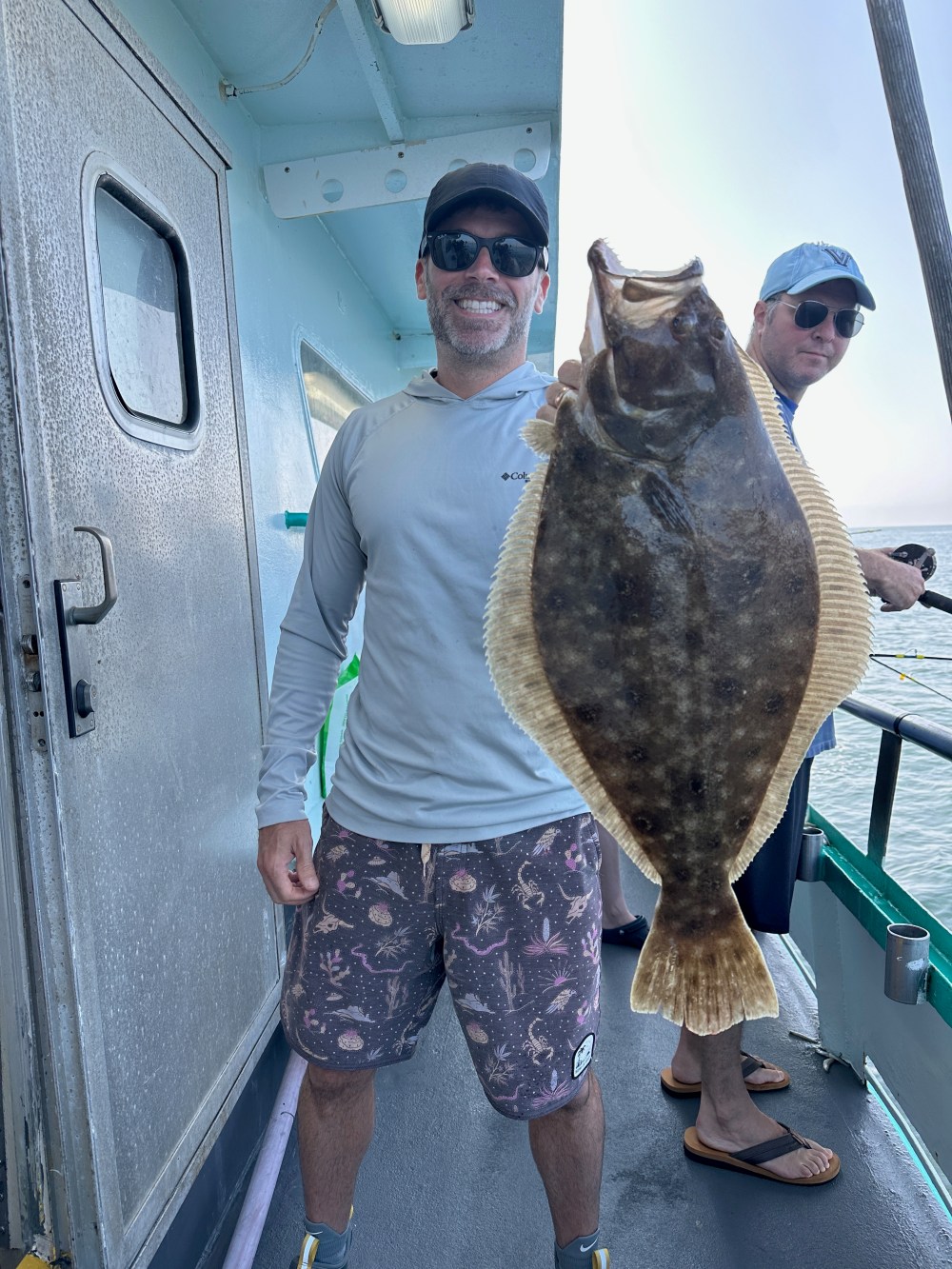 Man on a boat holding a large fish, smiling in sunglasses, another man in background.