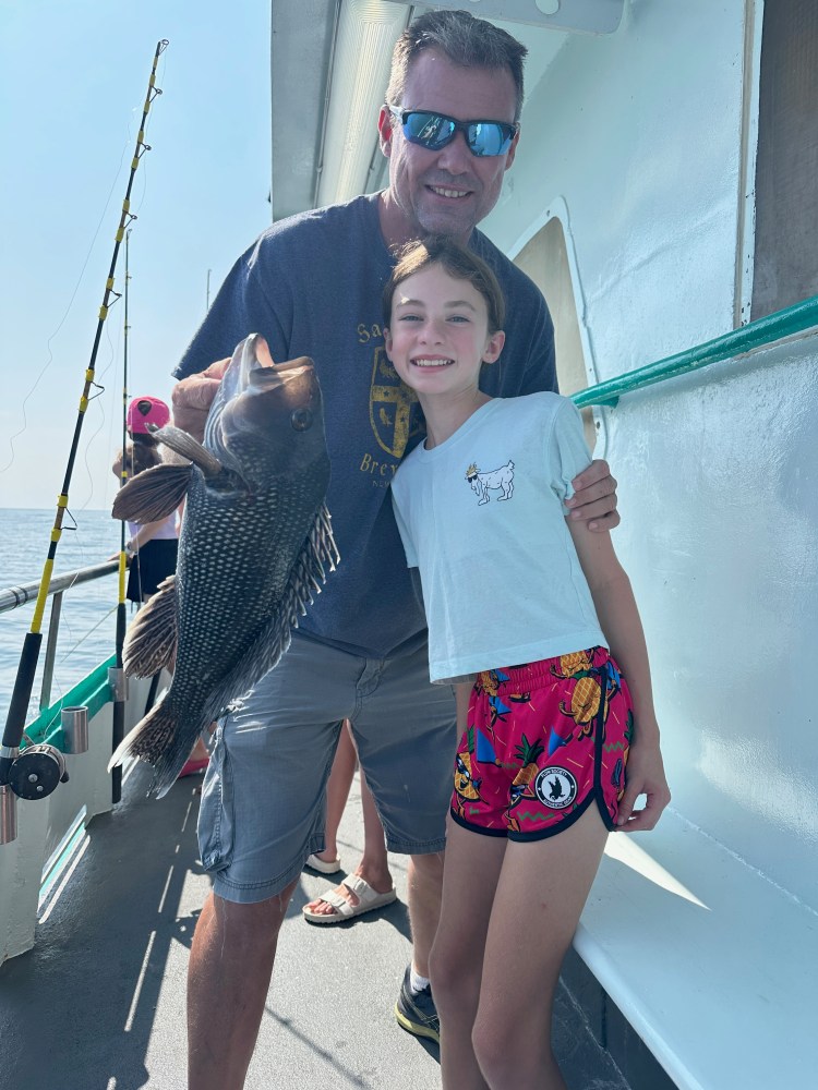 Two people smiling on a boat, holding a large fish with a fishing rod visible.