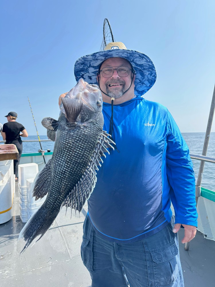 Man in blue shirt and hat holding a large fish on a boat, smiling at the camera.