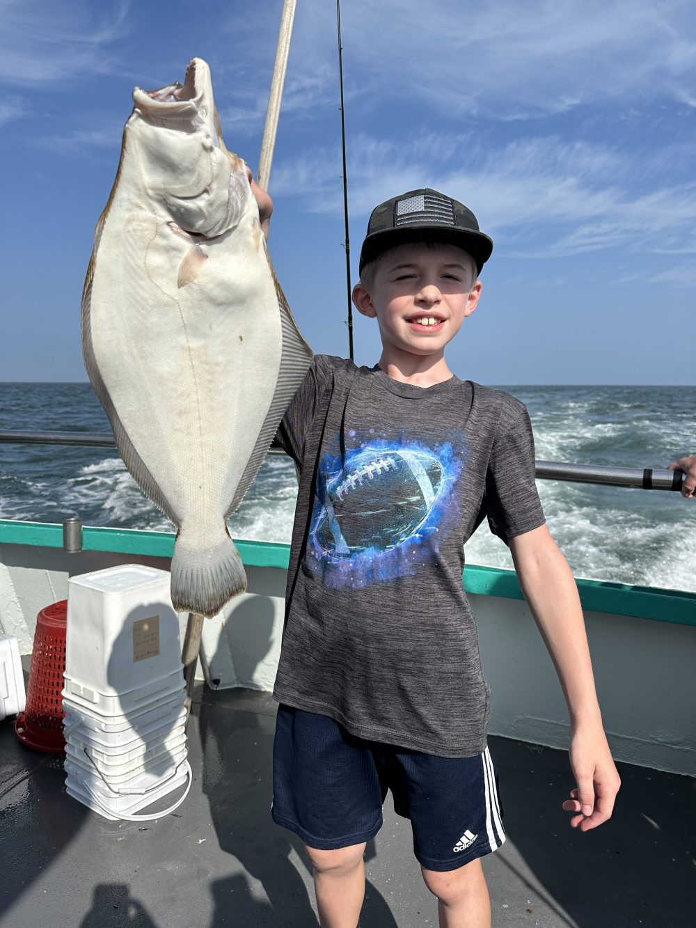Boy holding a large fish on a boat, ocean and sky in background.