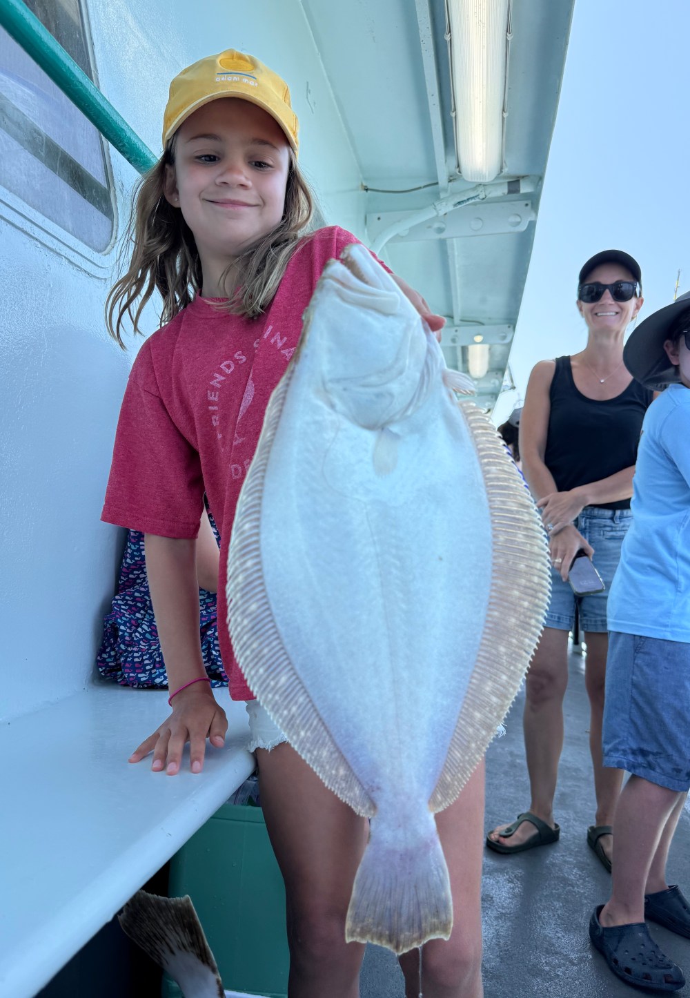 Child on a boat holding a large flat fish, with two people smiling in the background.