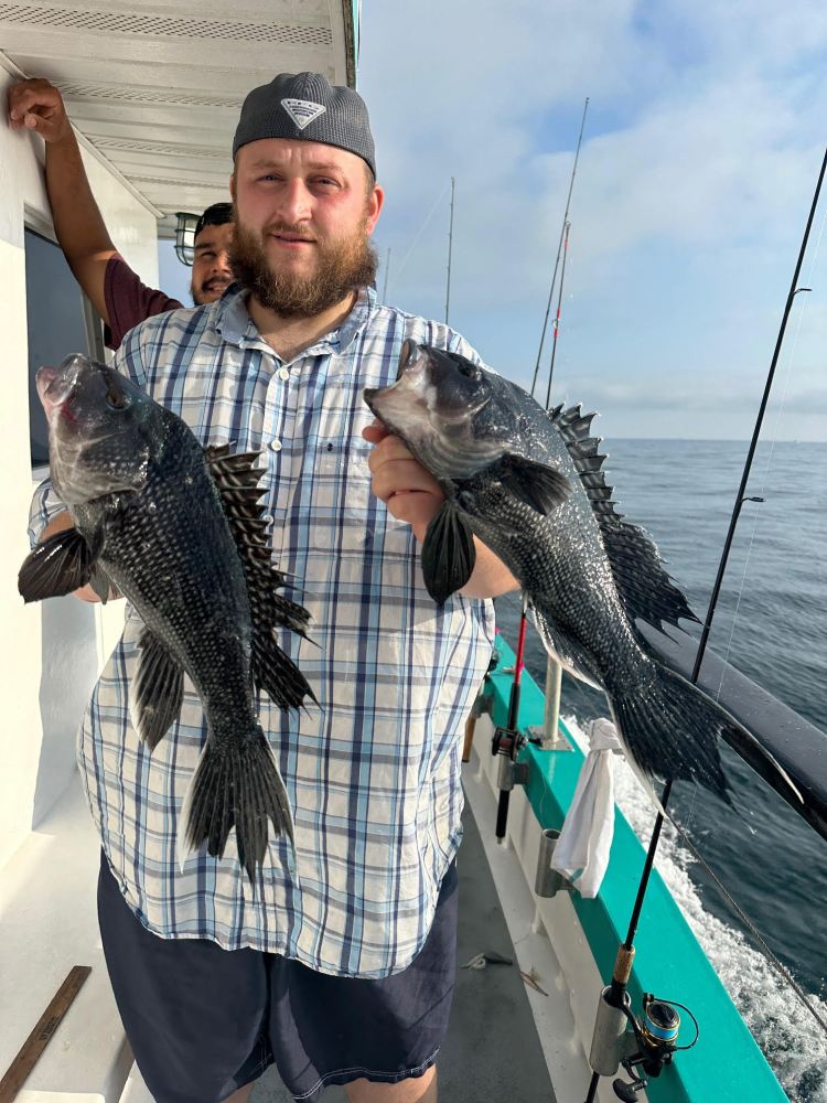 Man holding two large fish on a boat with fishing rods and ocean in the background.