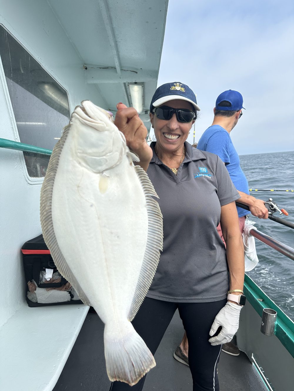 Smiling woman on a boat holding a large fish, wearing sunglasses and a cap.