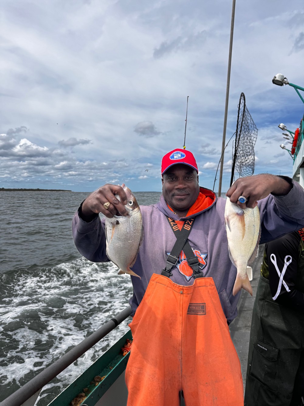 Man on a boat holding two fish, wearing orange overalls and red cap, with water and cloudy sky in the background.