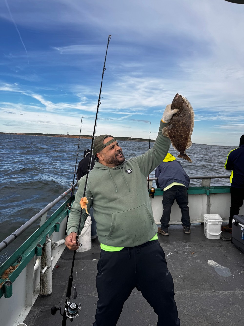 Man in hoodie holding fish and fishing rod on boat with ocean in background.