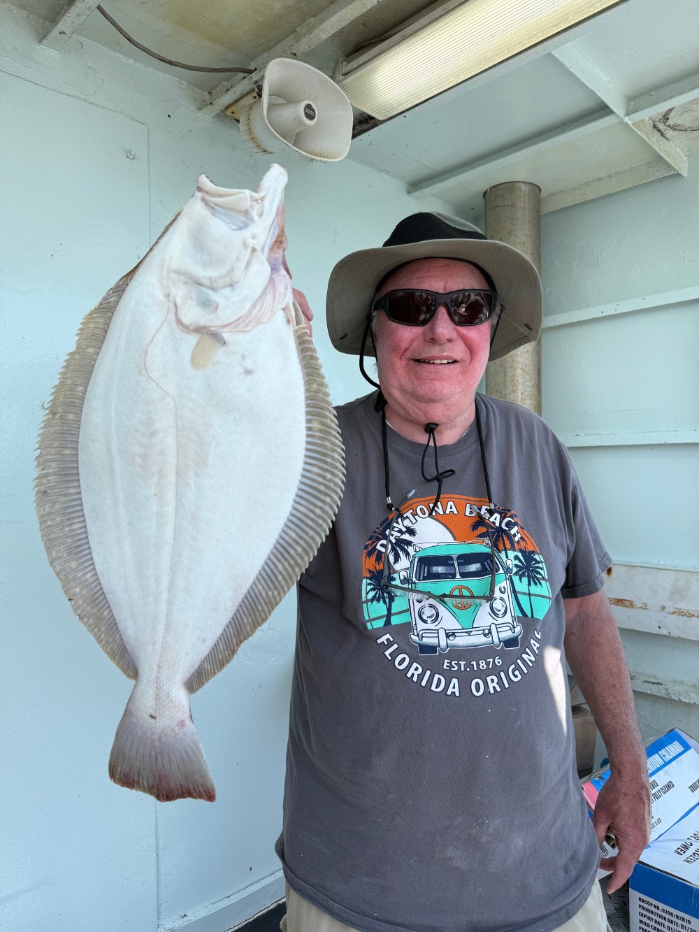 Man in hat holding a large flat fish on a boat, smiling at the camera.
