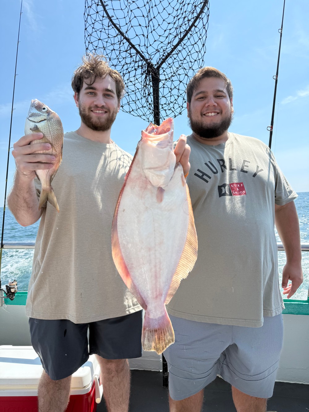 Two people smiling on a boat holding fish with a net and ocean in the background.