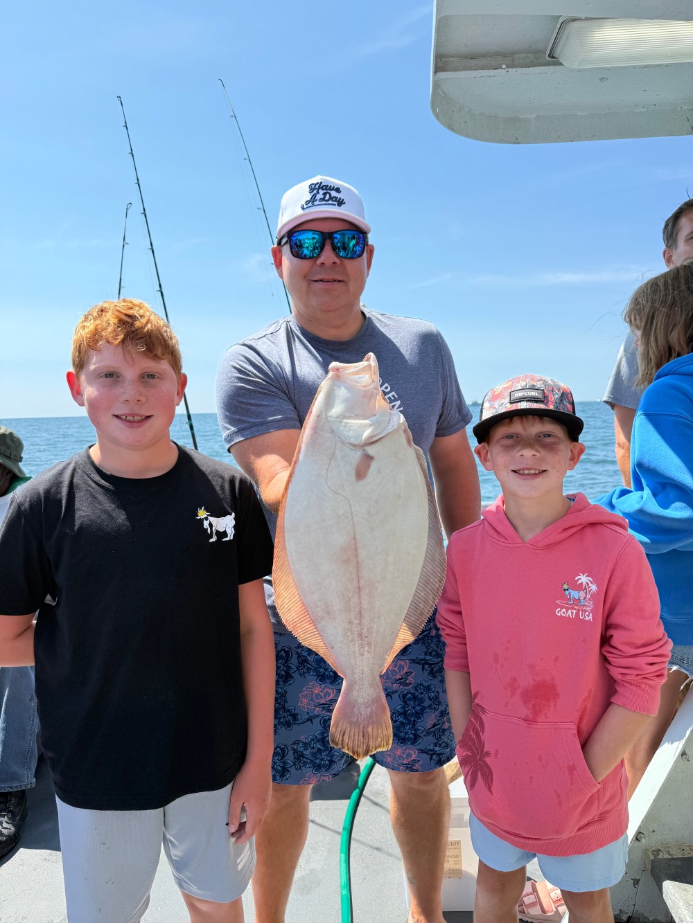 Two boys and a man hold a large fish on a boat under a clear blue sky.