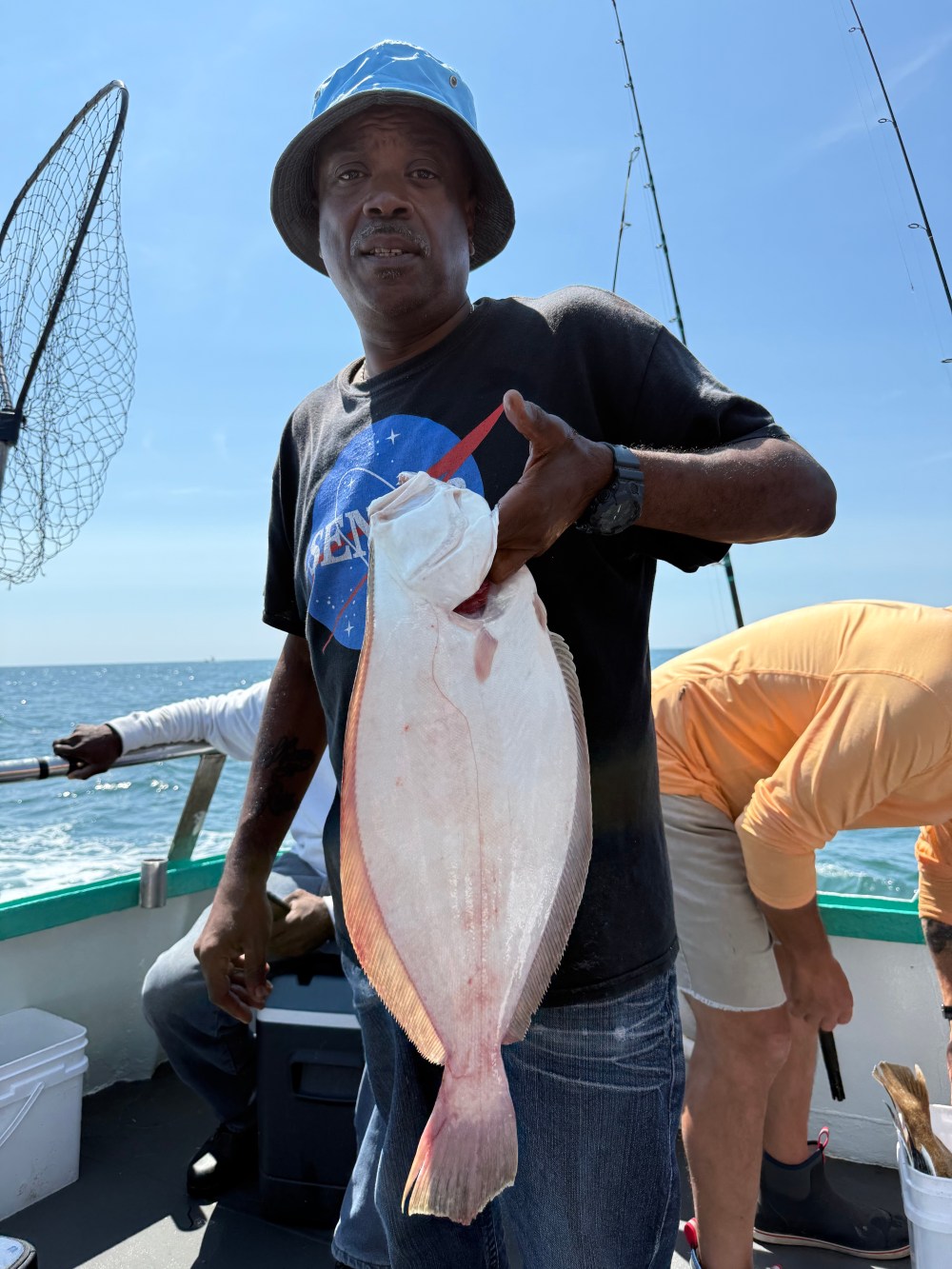 Man on boat holding a large flat fish, wearing a blue hat and dark shirt.