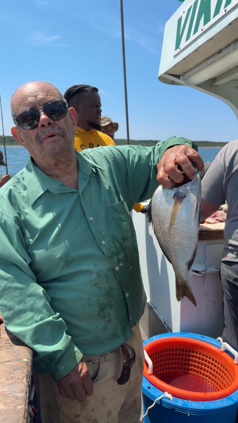Man holding a fish on a boat with others in the background under a clear sky.