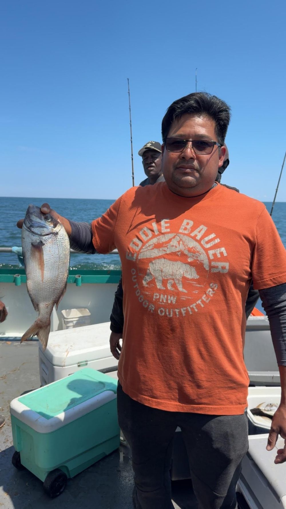 Man in orange shirt holding a fish on a boat with the ocean in the background.