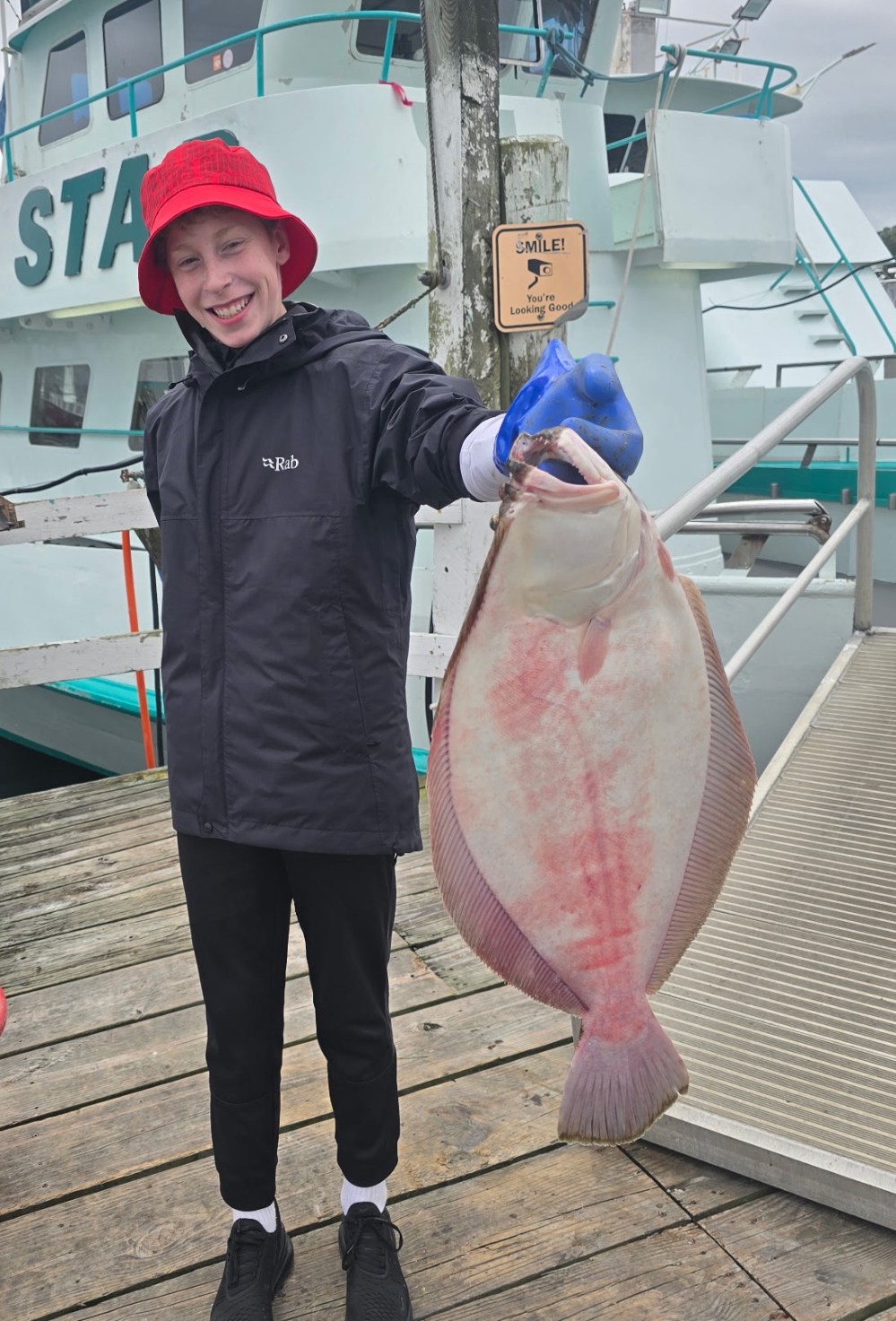 Person in red hat holding a large fish on a dock with a boat in the background.