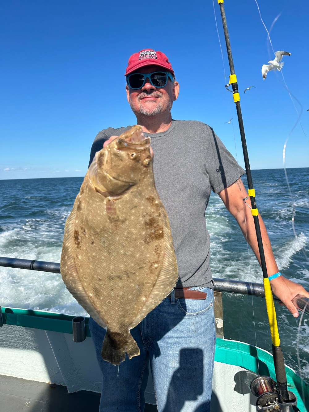 Person holding a large flatfish with fishing rod on a boat, ocean and sky in background.