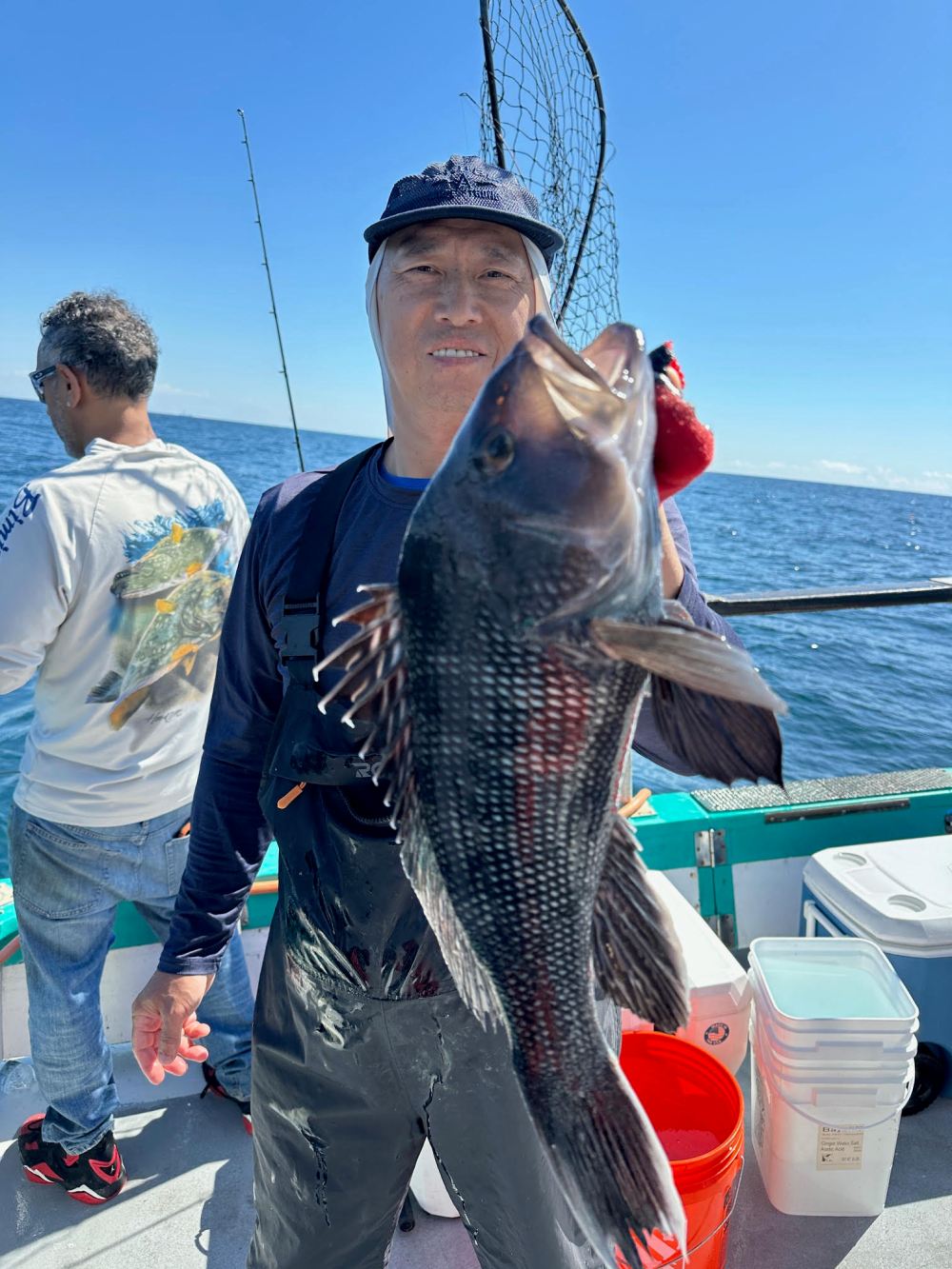 Person on a boat holds a large fish; another person stands in the background, with the ocean visible.