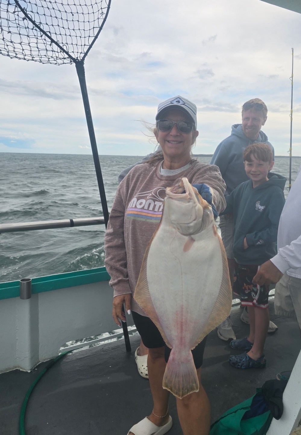 Woman holding a large flatfish on a boat with ocean and people in the background.
