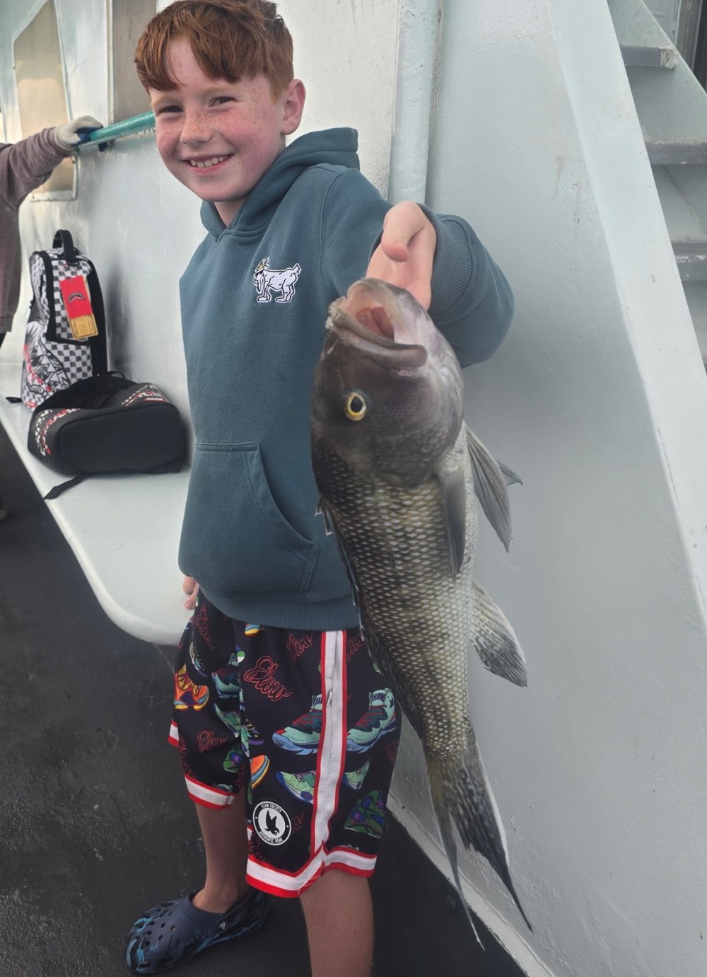 Child holding a fish and smiling on a boat, wearing a blue hoodie and patterned shorts.