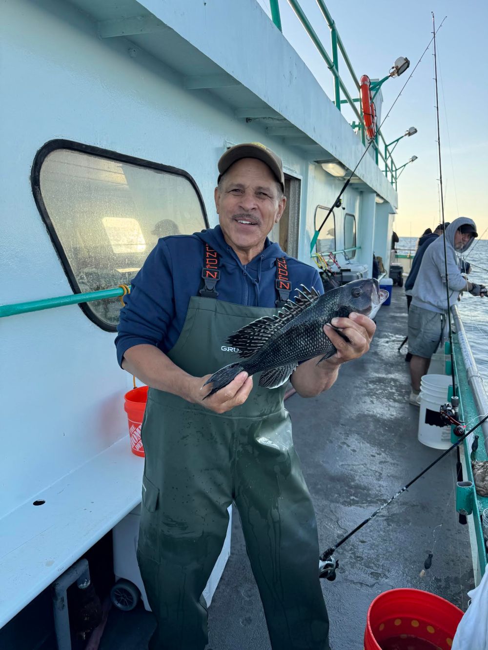 Man in waterproof gear holding a fish on a boat deck, with fishing rod and ocean in background.