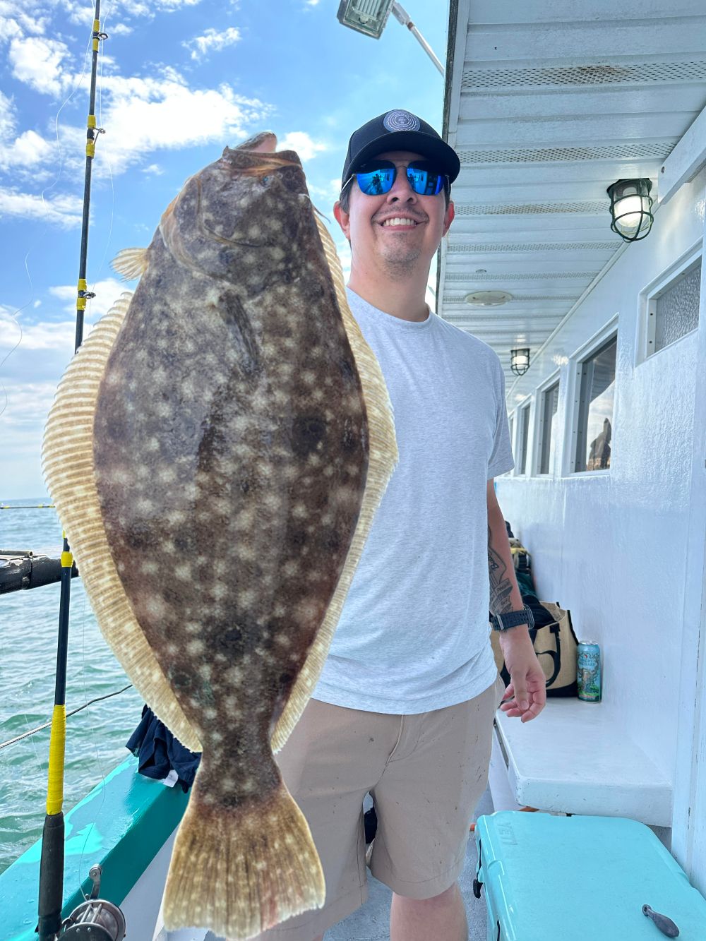 Person in sunglasses holding large flatfish on a boat under a clear sky.
