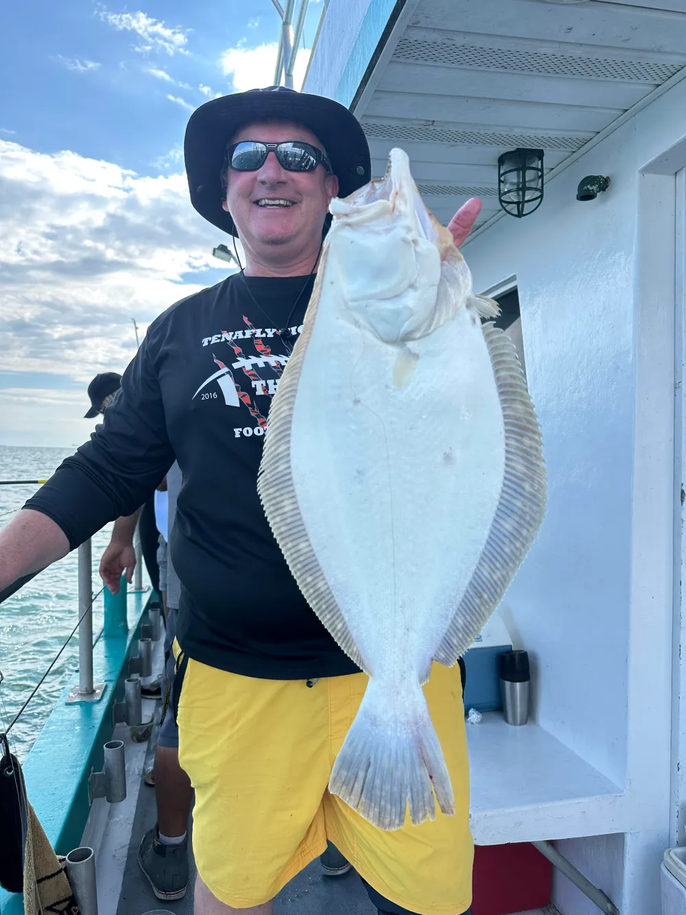 Person in hat and sunglasses holds a large fish on a boat under a cloudy sky.