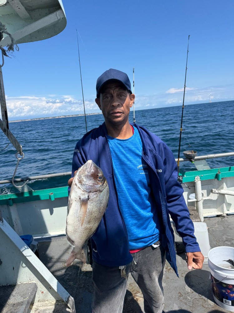 Person holding a fish on a boat, ocean in the background, wearing a blue cap and jacket.