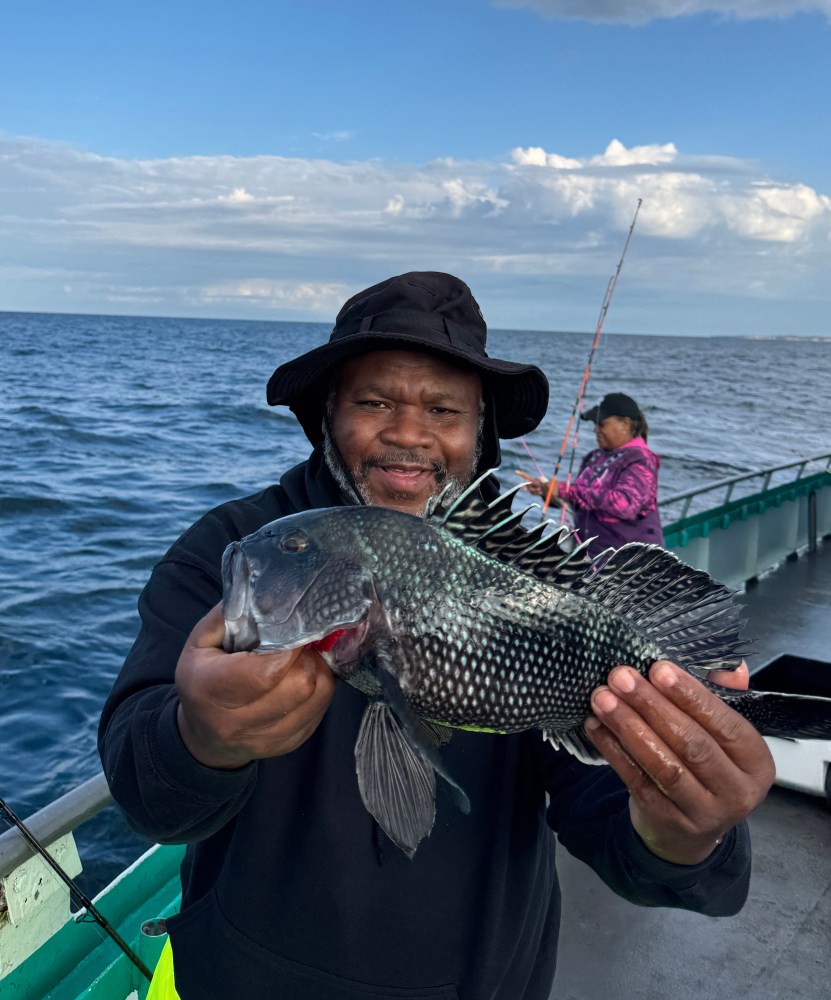 Smiling person on a boat holding a large fish, with ocean and another person in the background.