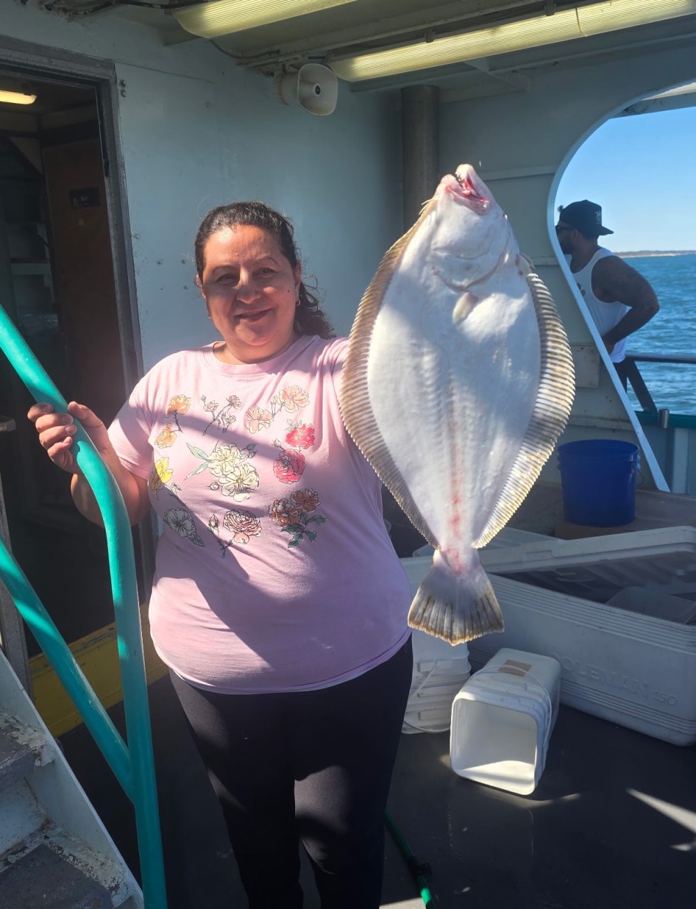 Person on a boat holding a large flatfish, smiling near stairs and open sea.