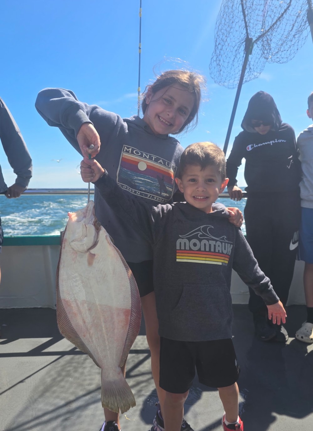 Two kids on a boat holding a large fish with a fishing net in the background.
