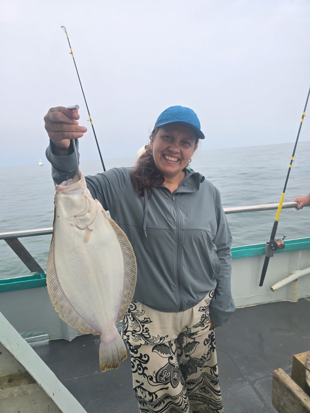 Smiling person holding a flat fish on a boat with fishing rods in the background.