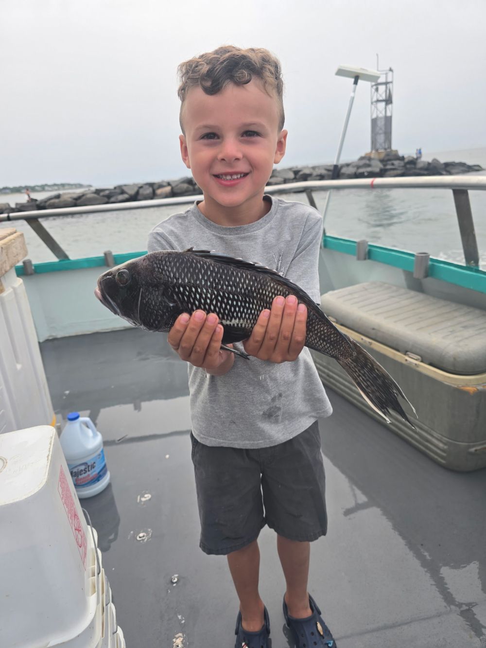 Child on boat holding a fish with a smile, ocean and rocky breakwater visible in the background.