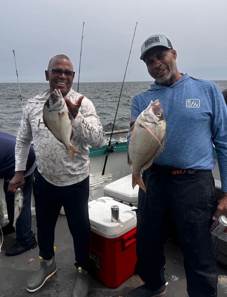 Two men smiling on a boat, each holding a fish, with a cloudy sea in the background.