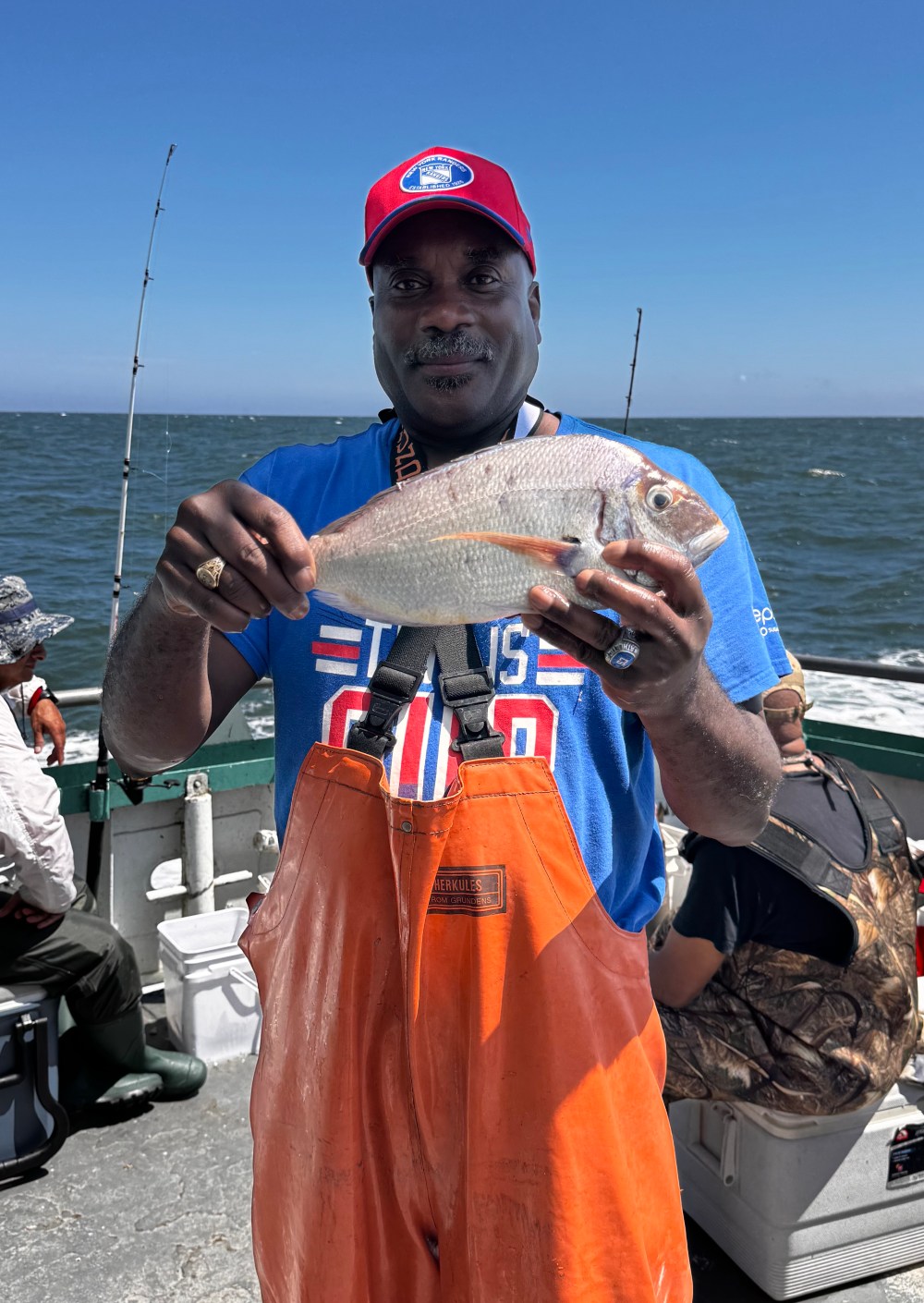 Man in orange overalls holding a fish on a boat with ocean background.