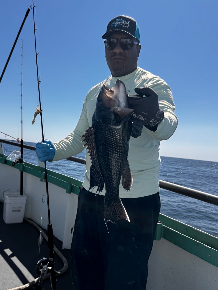 Man in a cap holding a large fish on a boat with ocean backdrop.