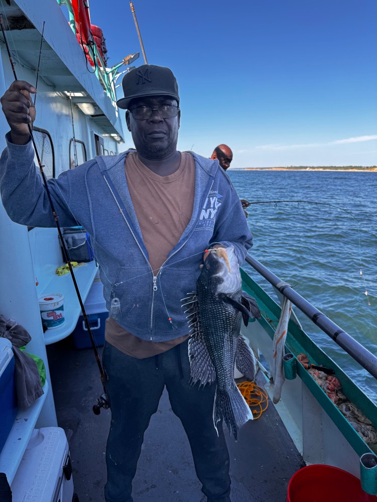 Man holding a large fish on a boat, wearing a cap and jacket, with water and sky in the background.