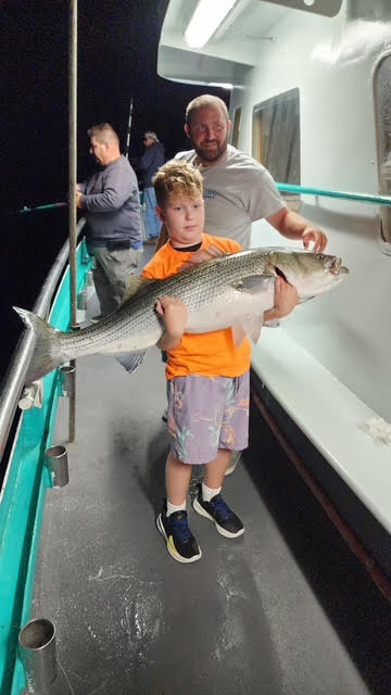 Boy proudly holds large fish on a boat at night, with two adults in the background.