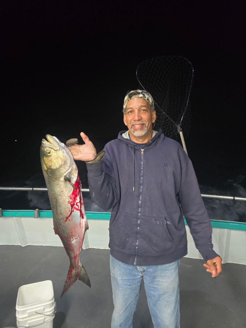 Man in cap holding large fish with red markings on boat at night.