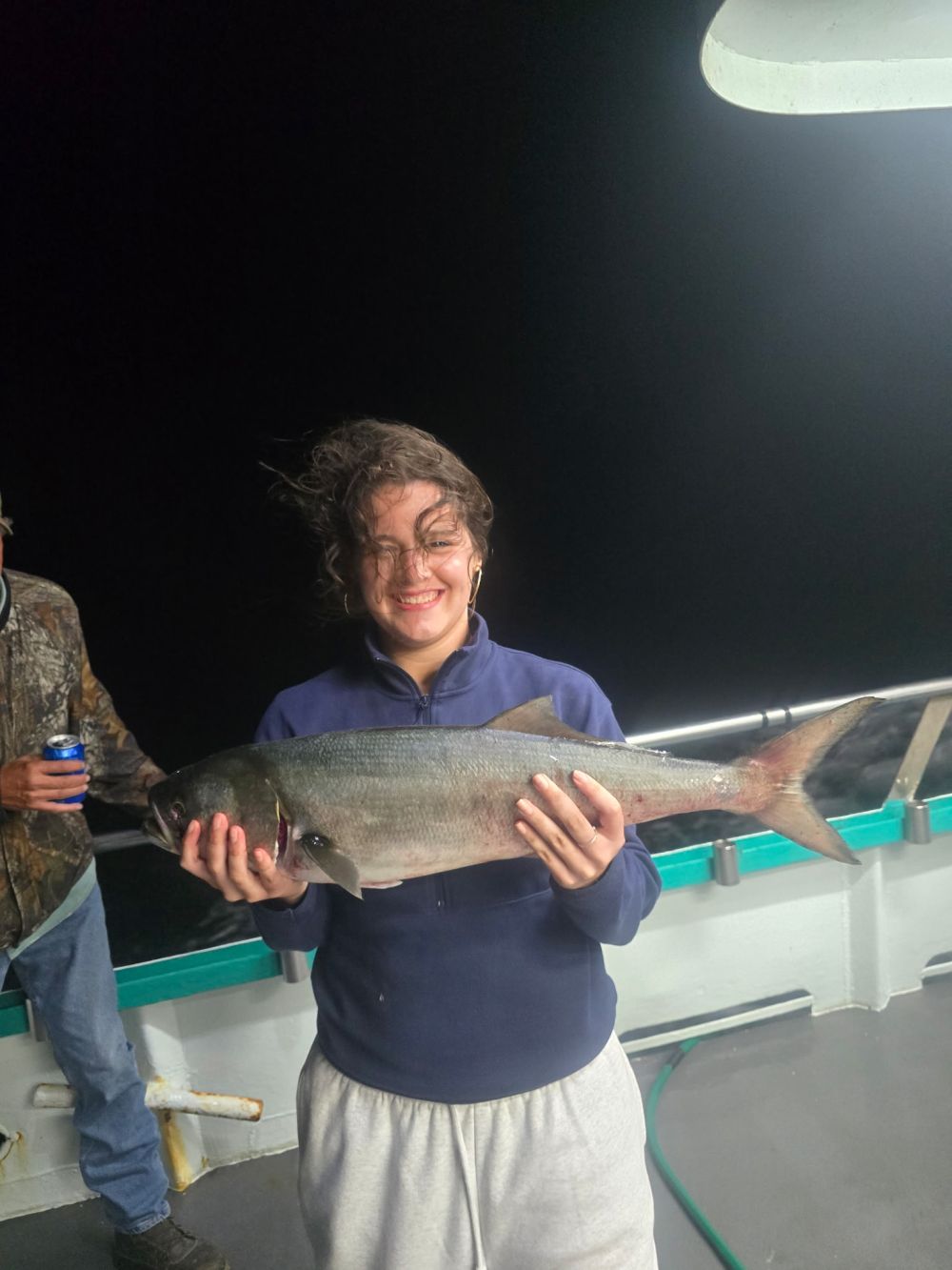 Person holding a large fish on a boat at night with windblown hair.
