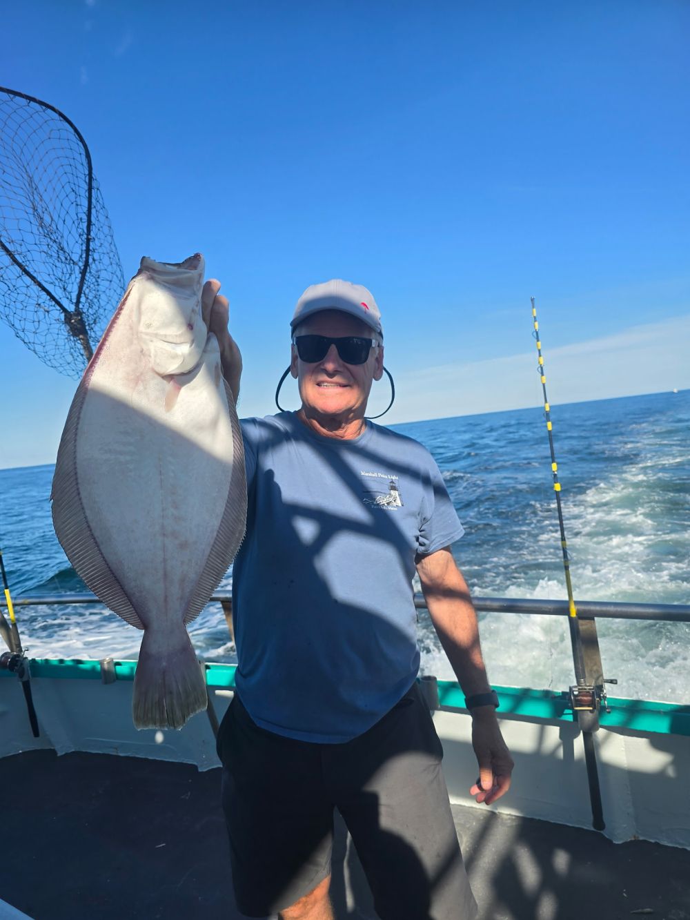 Man on boat holding a large fish with ocean and fishing rods in the background.